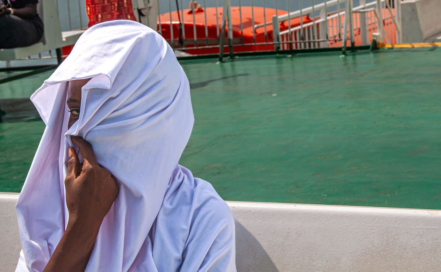 Person covering their face with a white cloth while sitting near green water on a boat.