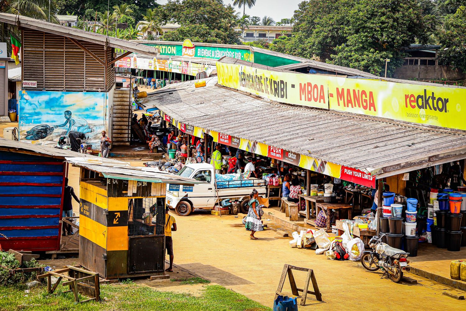 A busy outdoor market with colorful storefronts and signs including Manga and Reaktor on a dirt road.