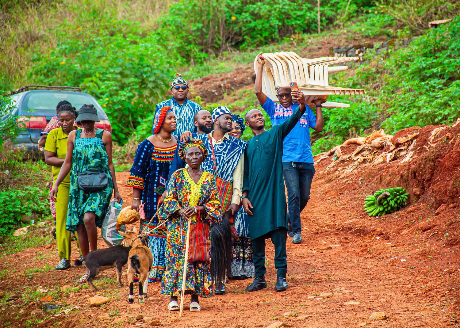 A group of people in colorful traditional African attire walking along a red dirt path in a lush green landscape.