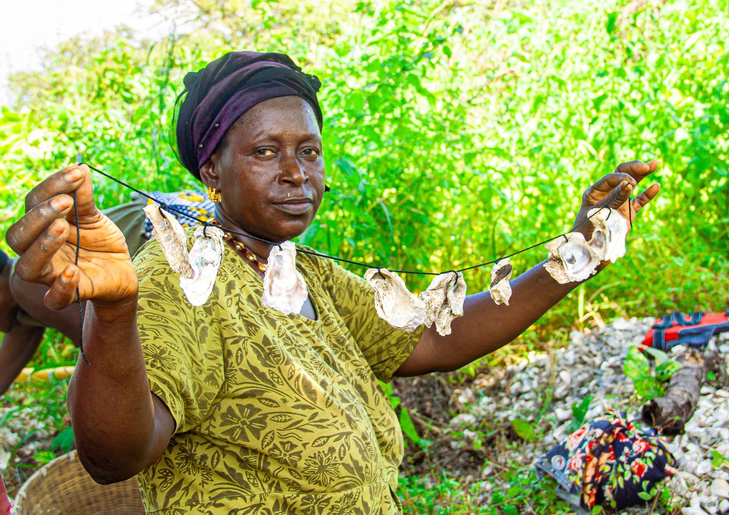 An African woman in olive clothing holds up a string of dried fish outdoors among green vegetation.