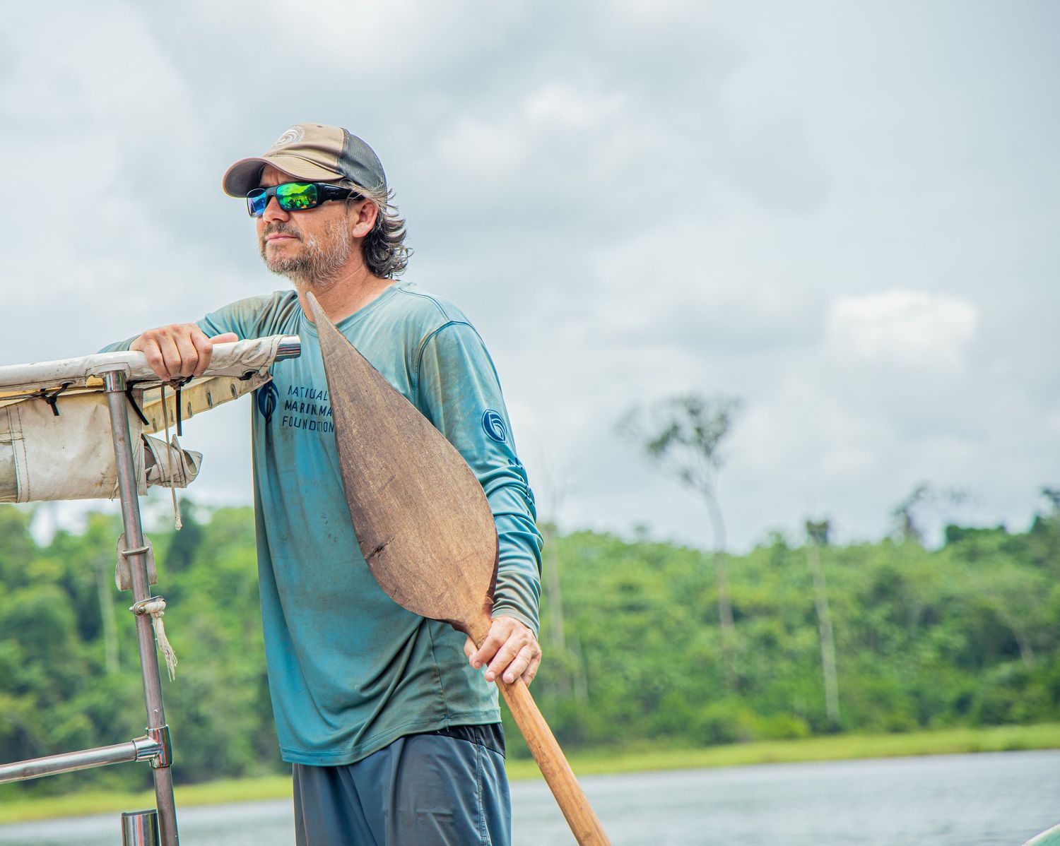 Man holding a wooden paddle on a boat with a river and green forest in the background.
