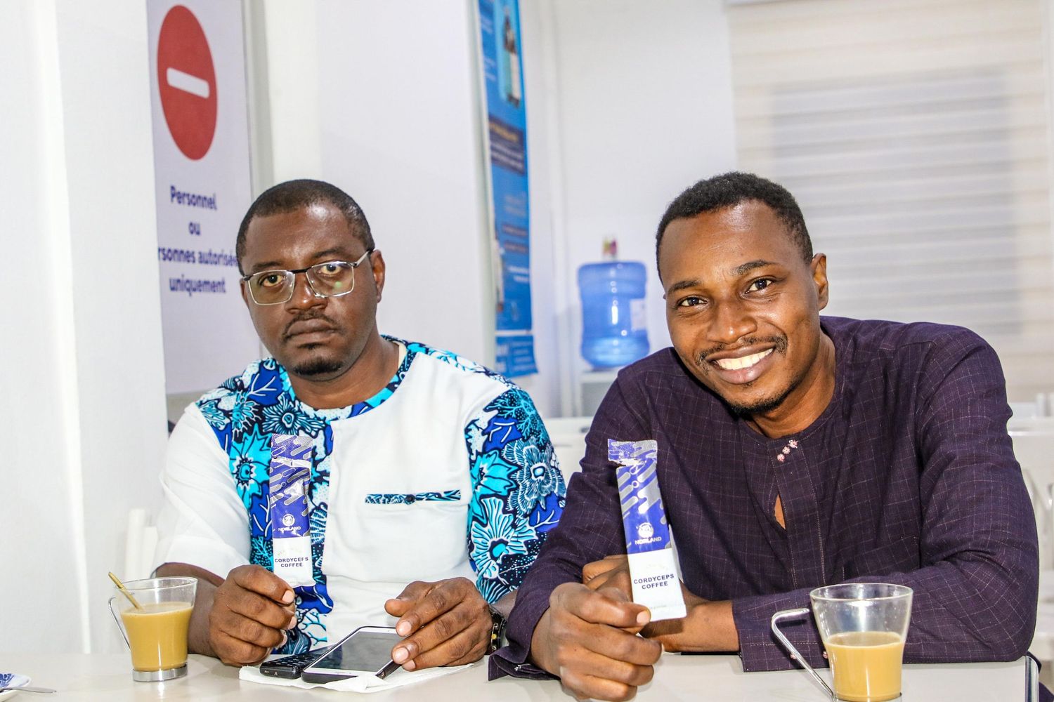 Two men smile at the camera while seated at a table, one holding a small blue product box.