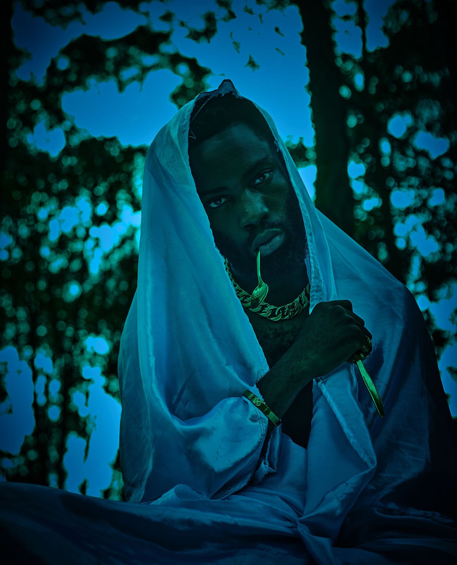 Man draped in white cloth with gold chain posing under blue-lit forest trees.