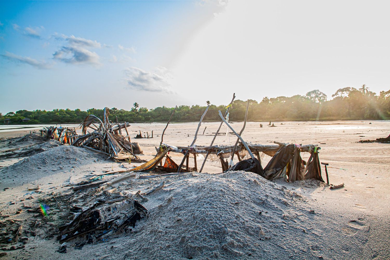 Abandoned wooden structure on a white sandy beach at sunset with lush trees behind.