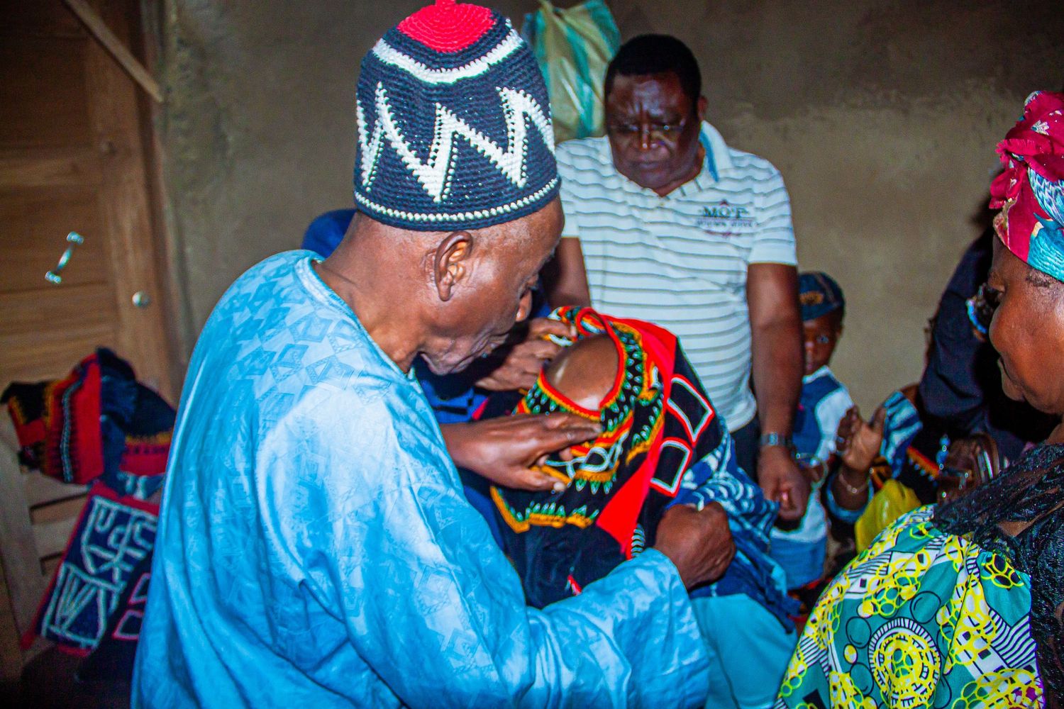 An elder in blue boubou examines colorful traditional African beaded regalia indoors with observers nearby.