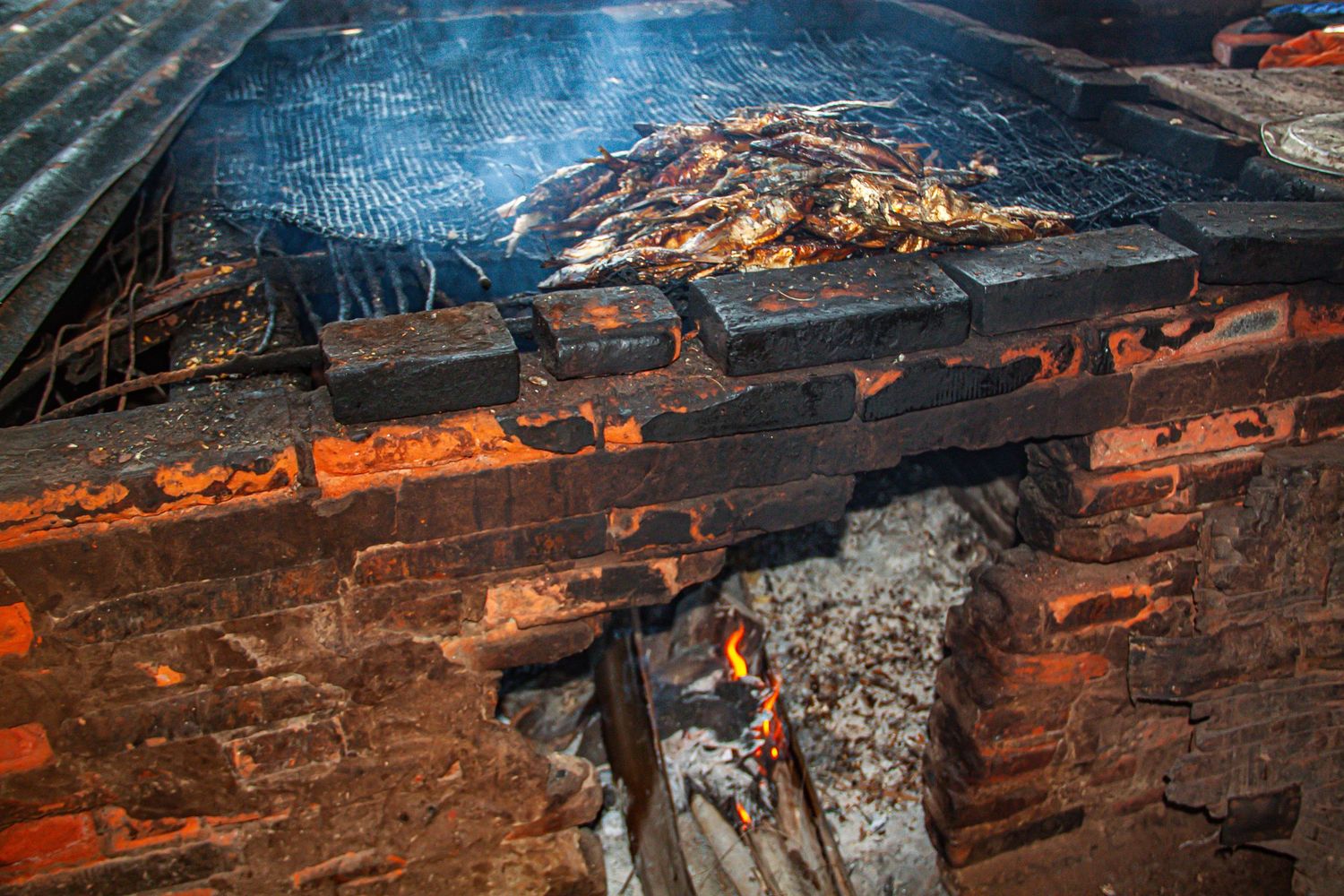 Fish smoking on a brick wood-fired grill with open flames below.