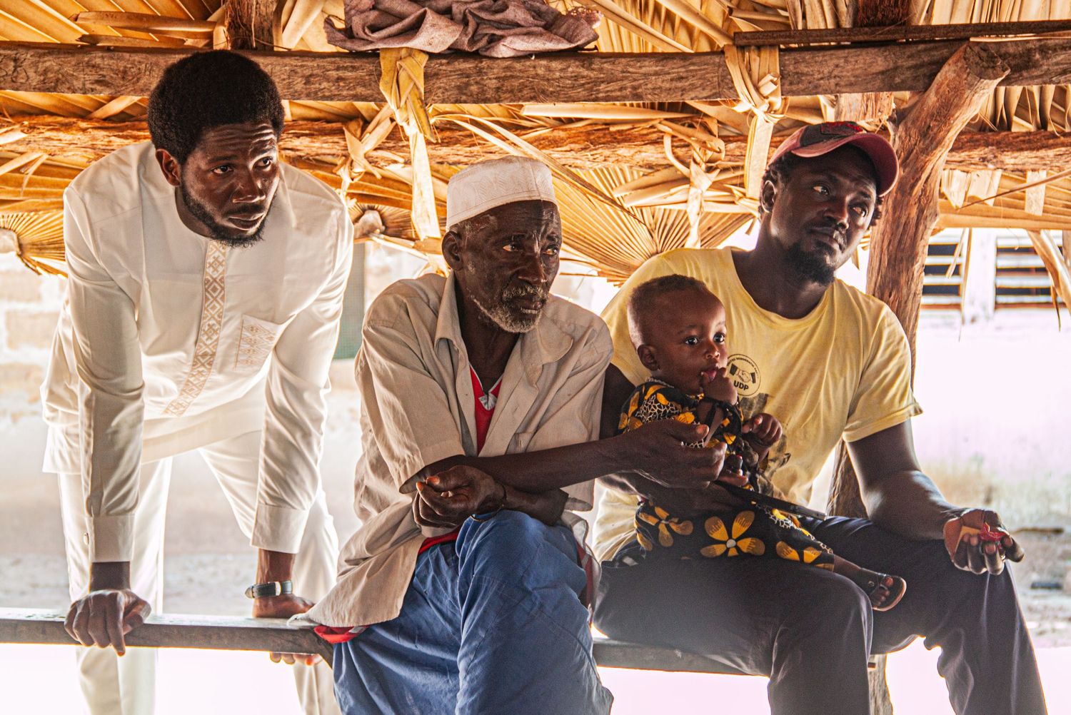 Three African men and a child sitting together under a thatched roof in a village gathering space.