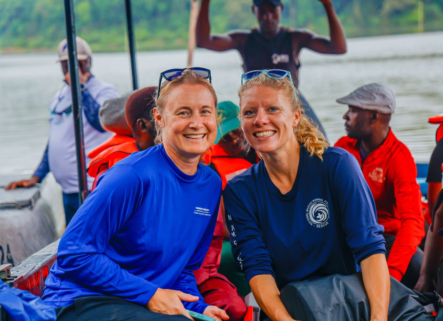 Two smiling women in blue shirts sitting on a boat on a calm river in a tropical location.