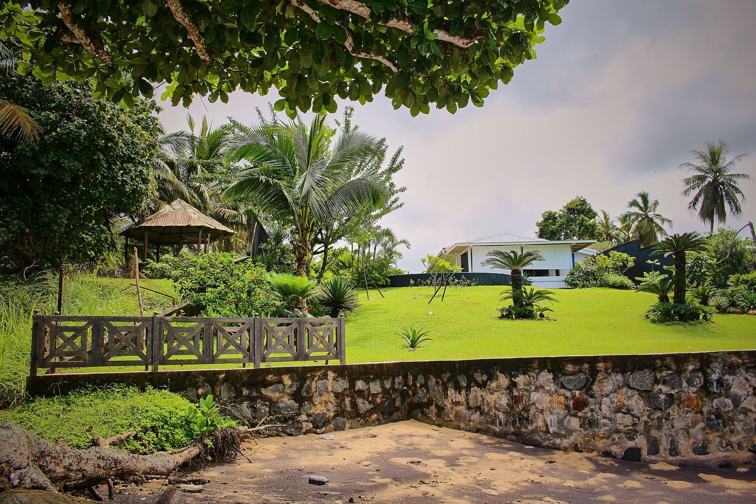 A lush tropical garden with manicured lawn, stone wall, wooden bridge, and palm trees under cloudy skies.