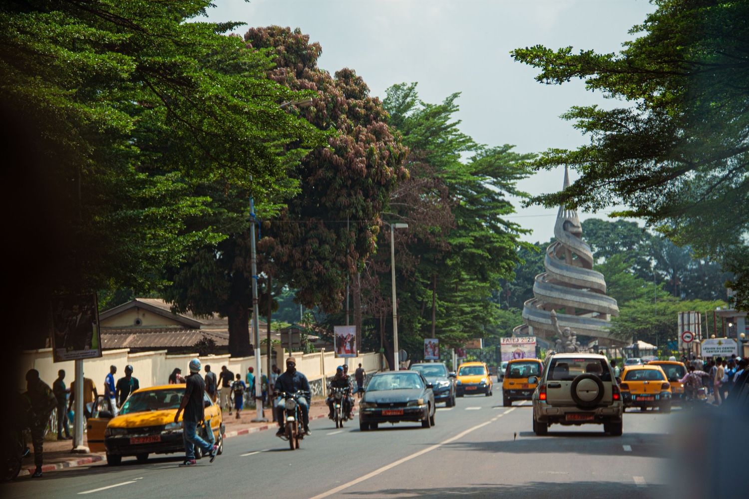 Busy African city boulevard lined with tall trees and colorful traffic in daylight.