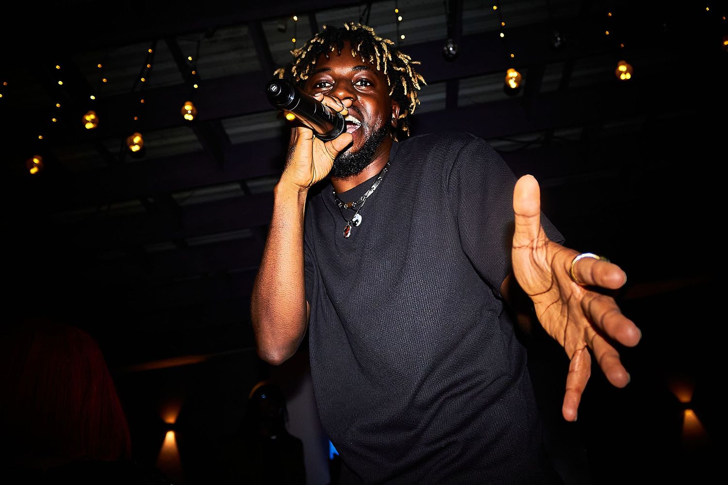 African male rapper performing energetically on stage with microphone under warm string lights at a music venue.