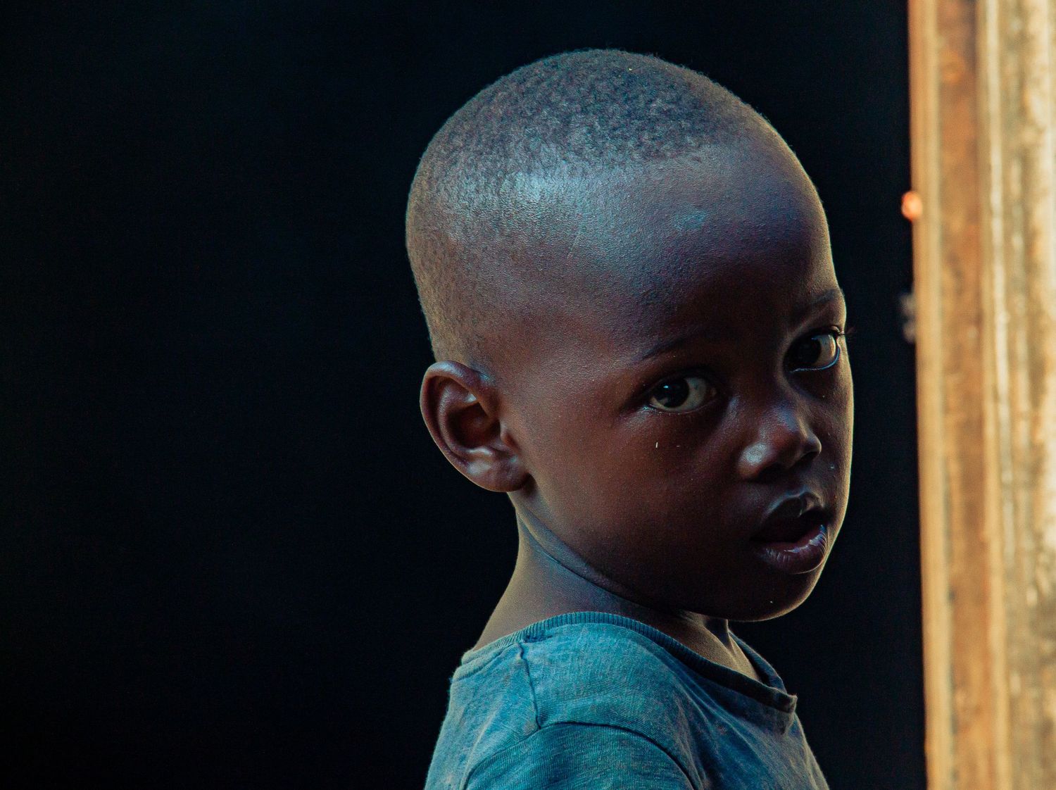 A young African boy in a teal shirt looks over his shoulder in dramatic lighting.