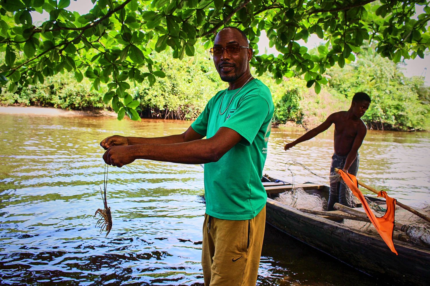Smiling African man holding a freshly caught fish on a wooden canoe in a lush tropical river setting.