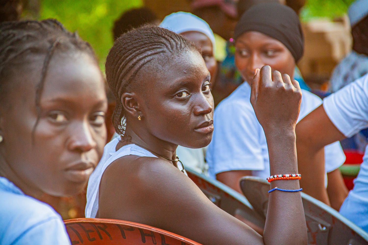 Young African woman with braided hair and bracelets seated attentively at an outdoor event.