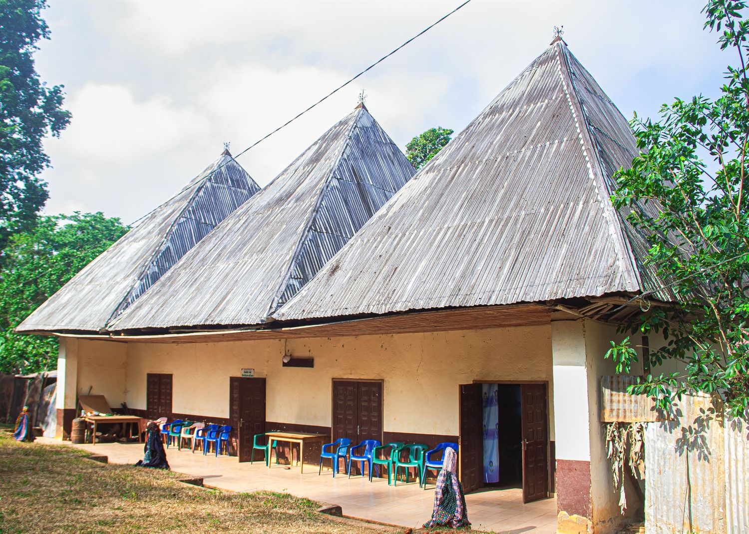Traditional African building with multiple thatched conical roofs surrounded by trees and outdoor seating.