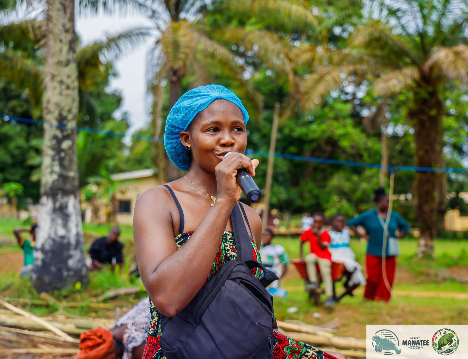 Woman speaking into microphone wearing blue headwrap at outdoor community event.