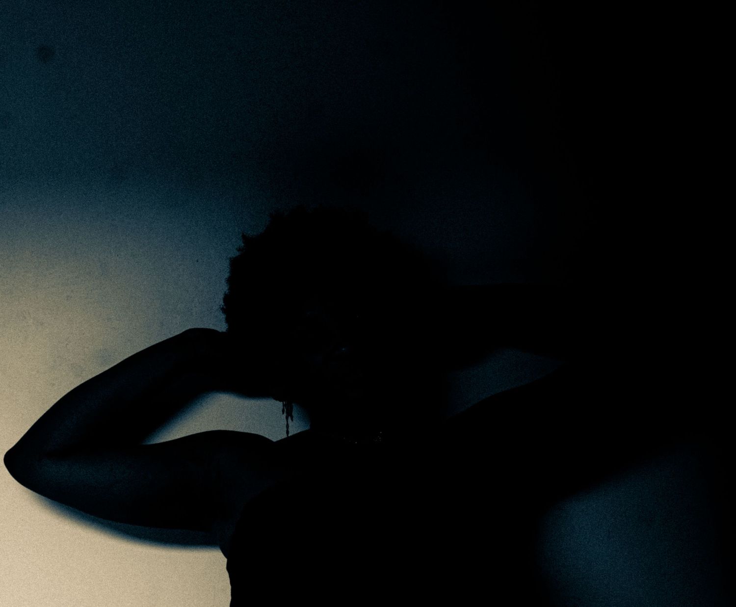 Dark silhouette of a person with hands raised to head against a dimly lit wall.