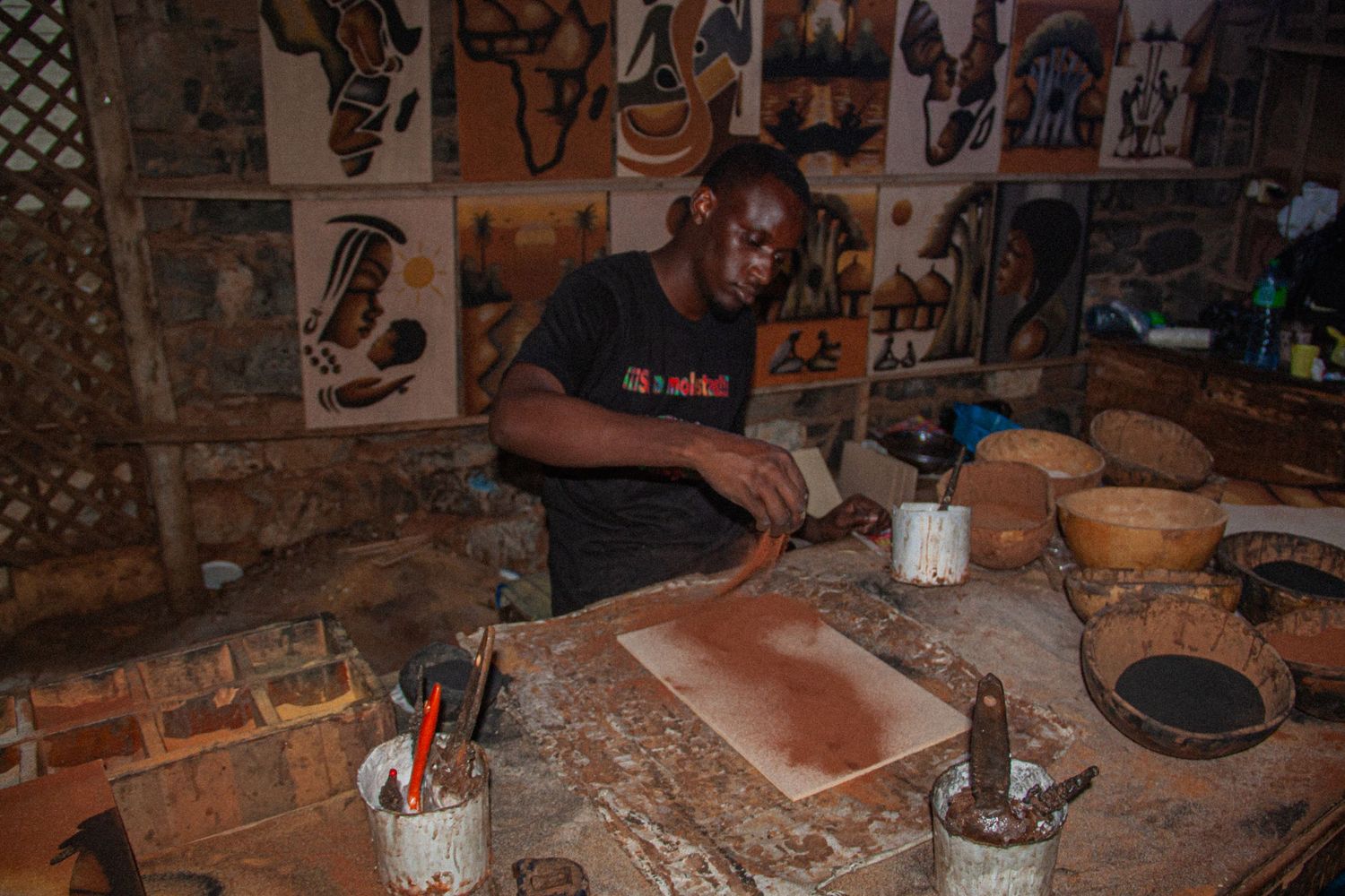 African craftsman working on traditional artwork in a rustic studio filled with cultural paintings.