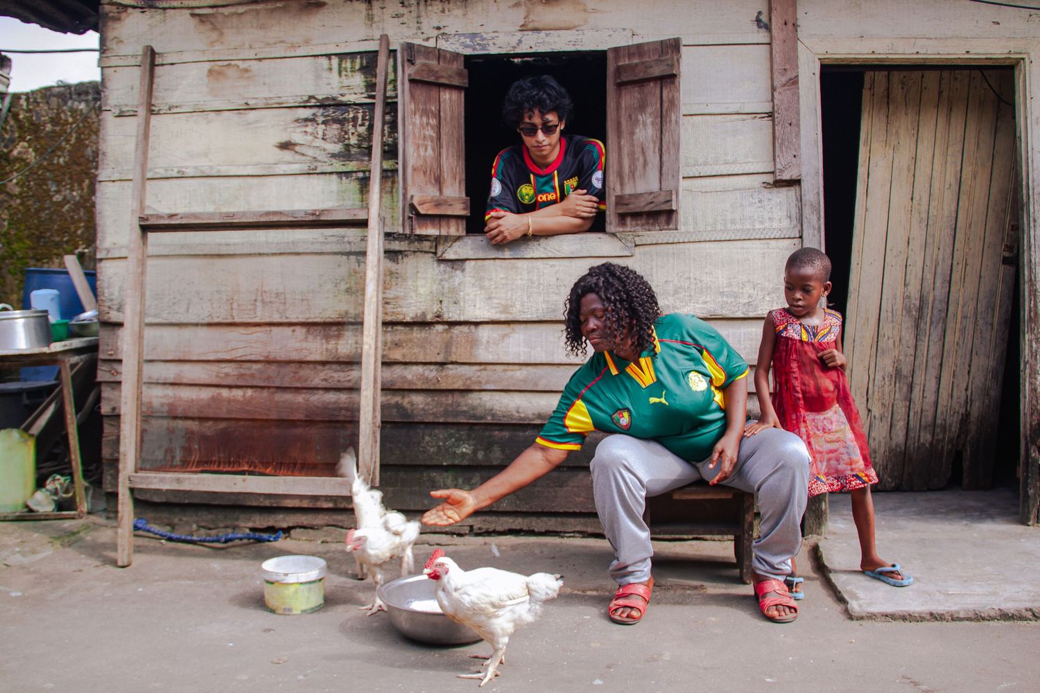 African family scene with person feeding white chickens outside a wooden house.