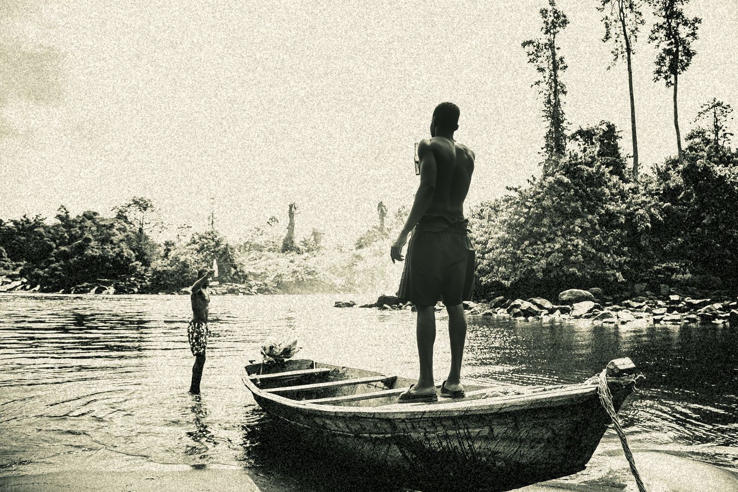 Vintage black and white photo of a man standing on a wooden canoe on a tropical river.