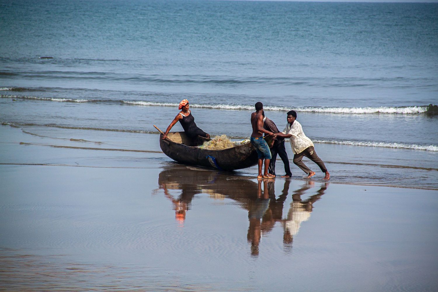 Three fishermen pushing a wooden boat into ocean waves on a sandy African beach.