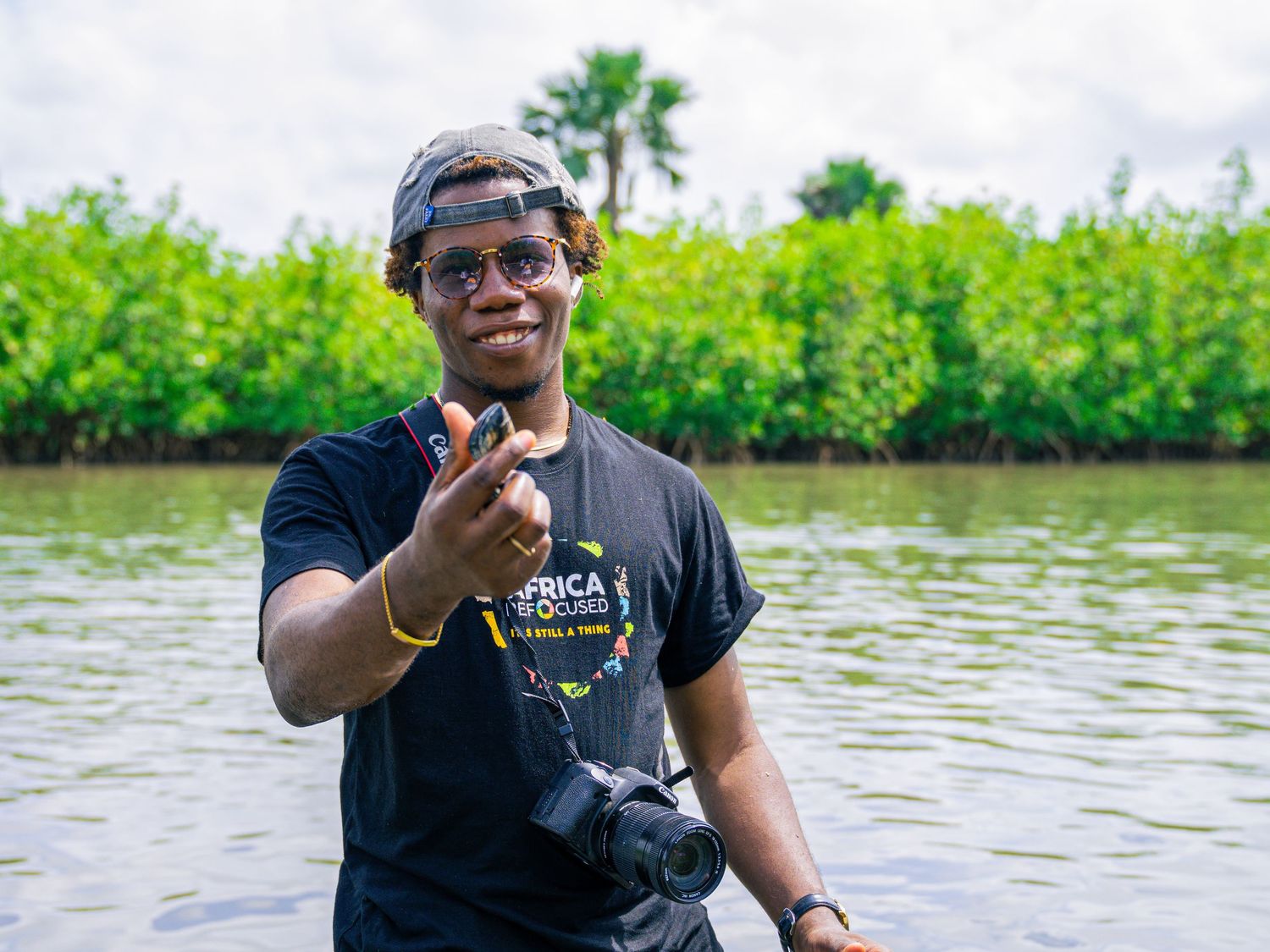 Smiling young man standing in shallow water wearing a black Africa Outdoors shirt holding a camera.