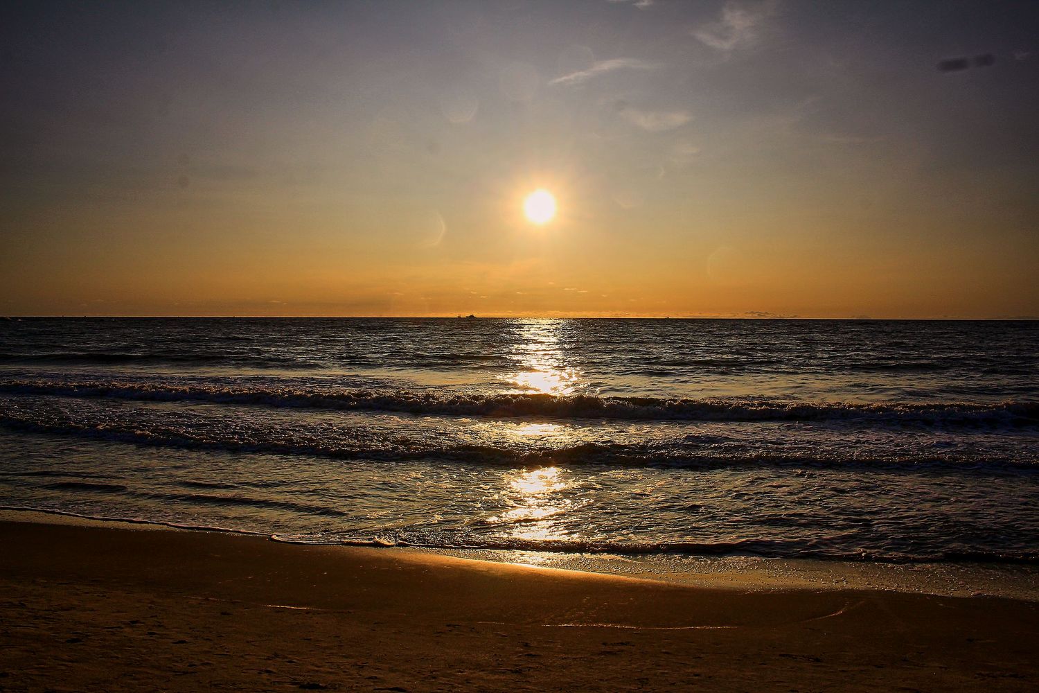 Golden beach sunset with gentle waves rolling onto sandy shore under a warm glowing orange sky.