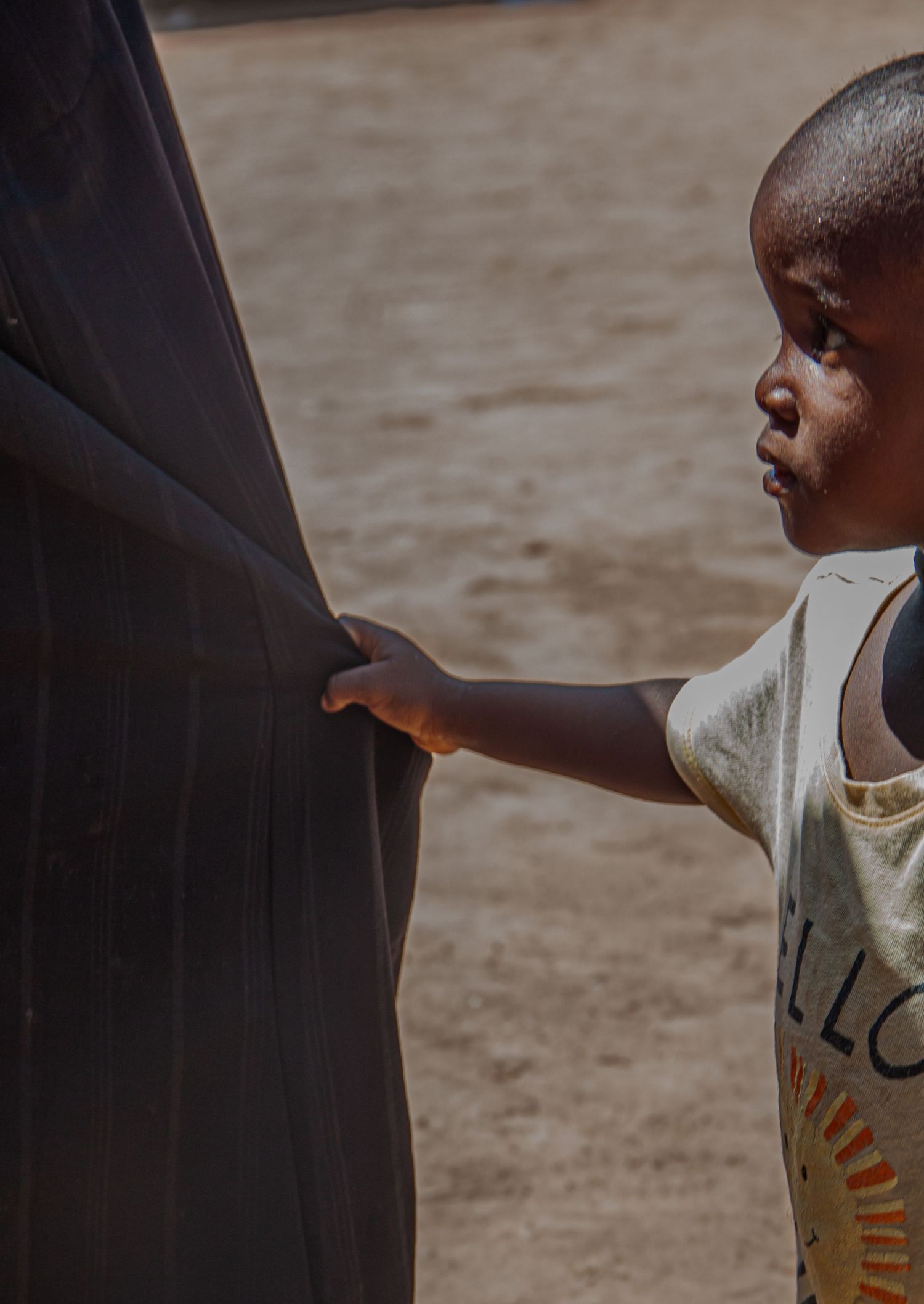 Young African child in a yellow shirt holding onto an adults dark robe in a sandy outdoor area.