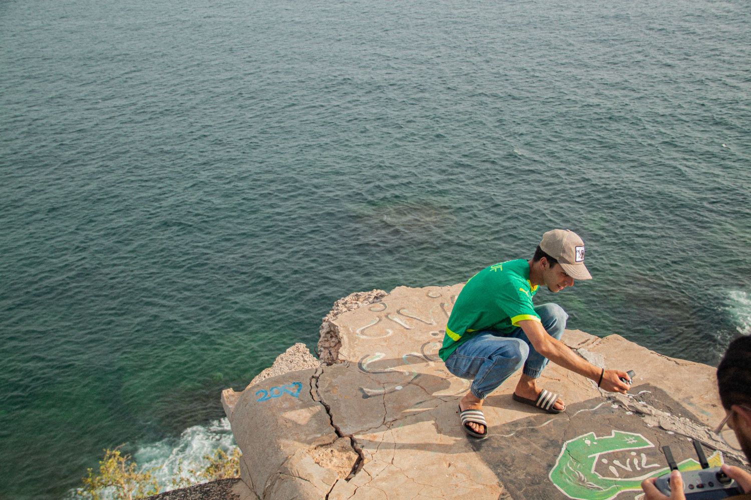 Person in green shirt crouching on rocky coastal cliff examining carvings above turquoise ocean water.