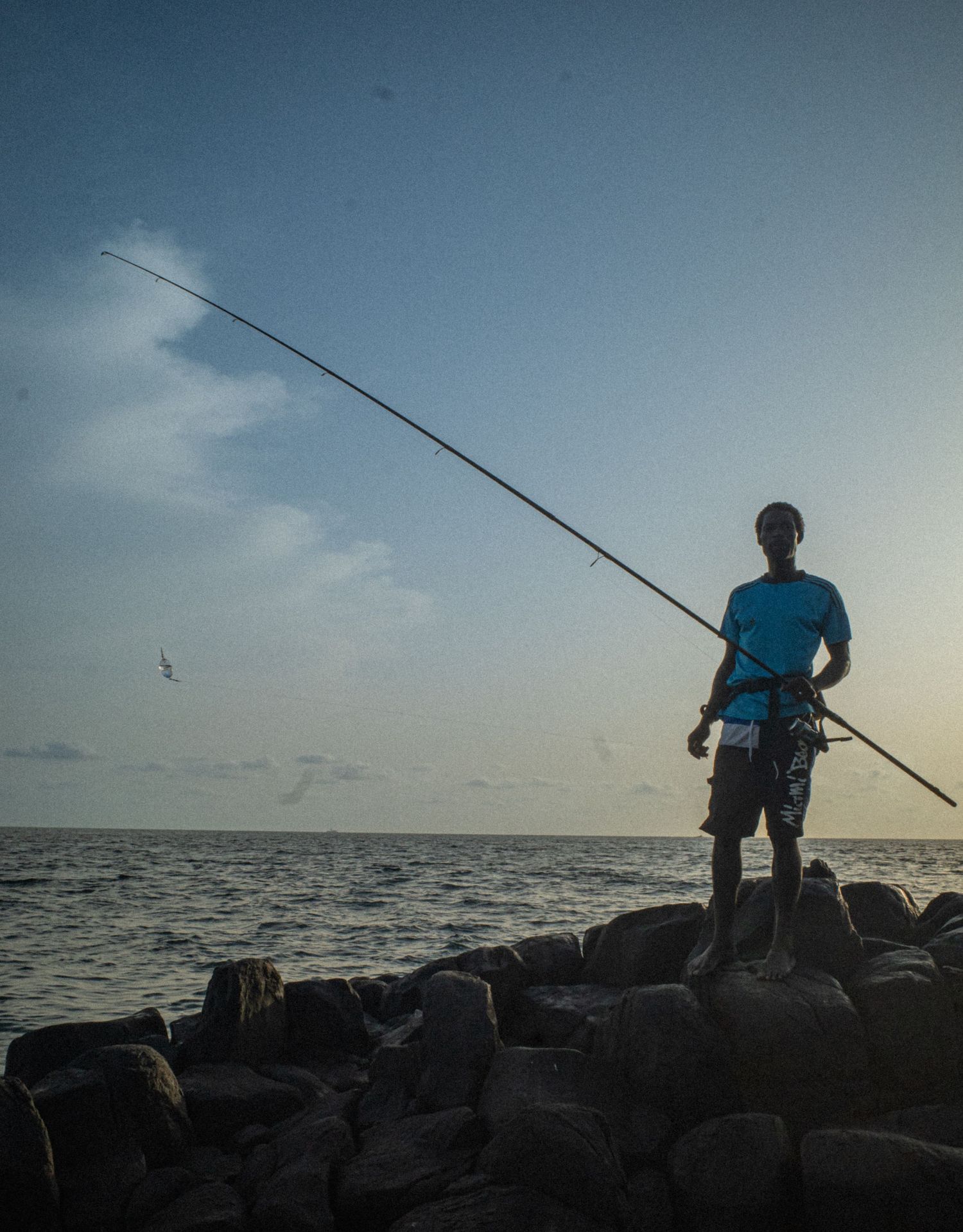 Silhouette of a young man fishing with a long rod standing on rocks at ocean sunset.