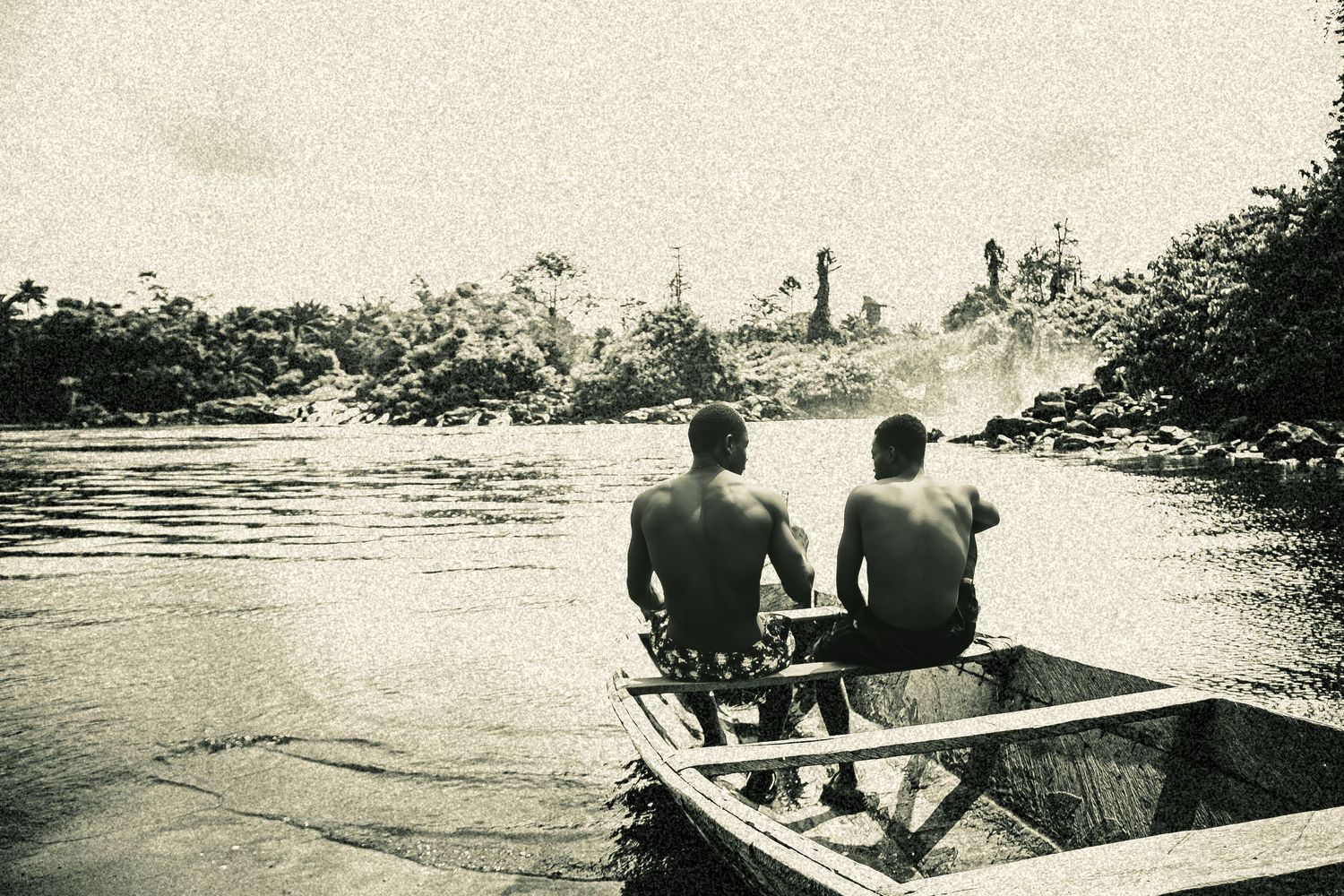Two men sitting in a wooden boat on a river surrounded by lush tropical vegetation.