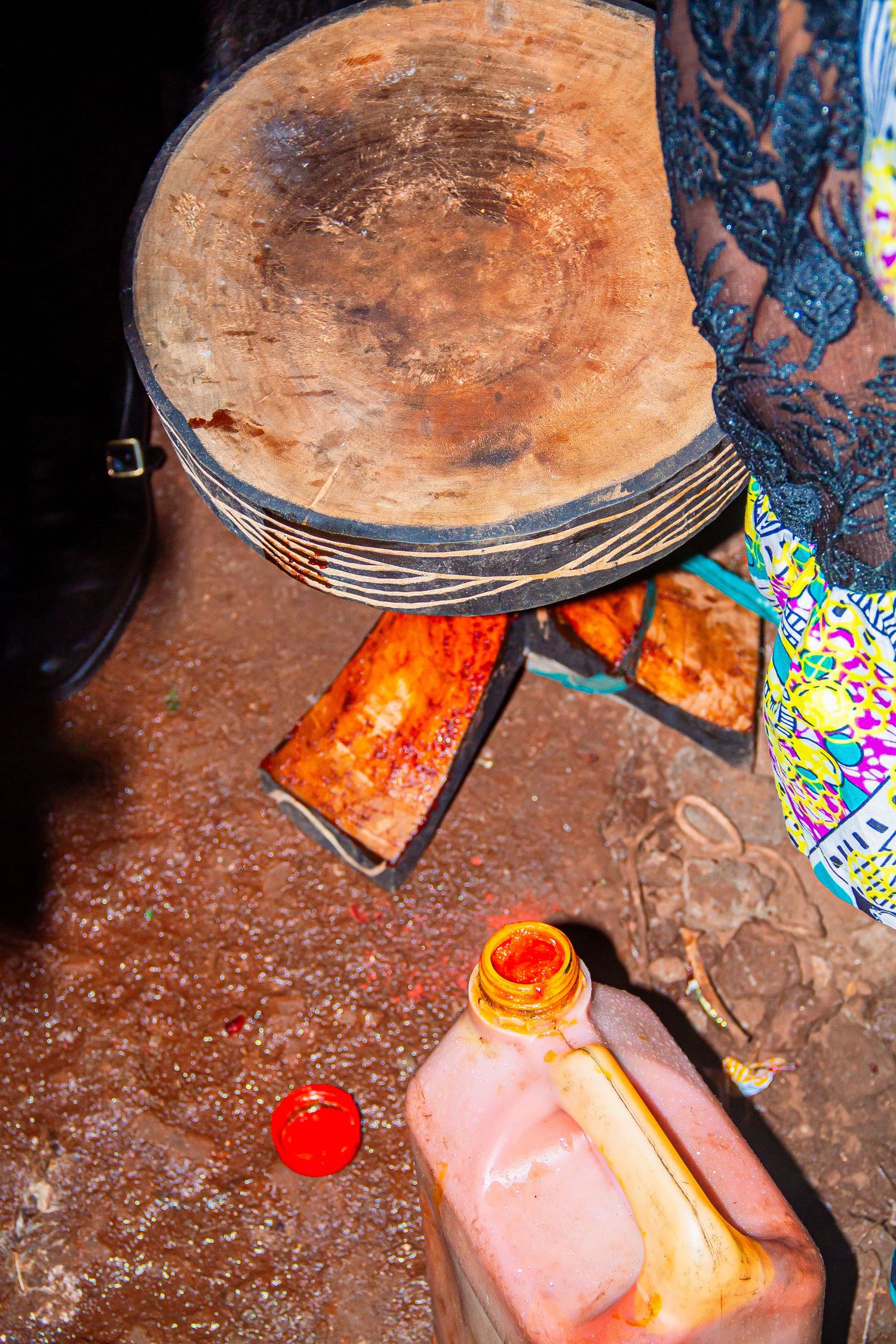 A close-up of a traditional wooden African drum on a stand with a red candle and plastic container below.