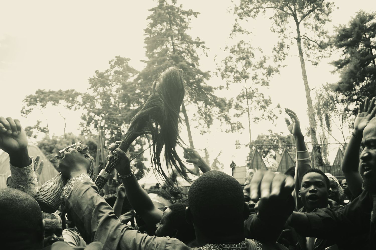 Black and white photo of a crowd at an outdoor festival, with a performer lifted above the audience among tall trees.