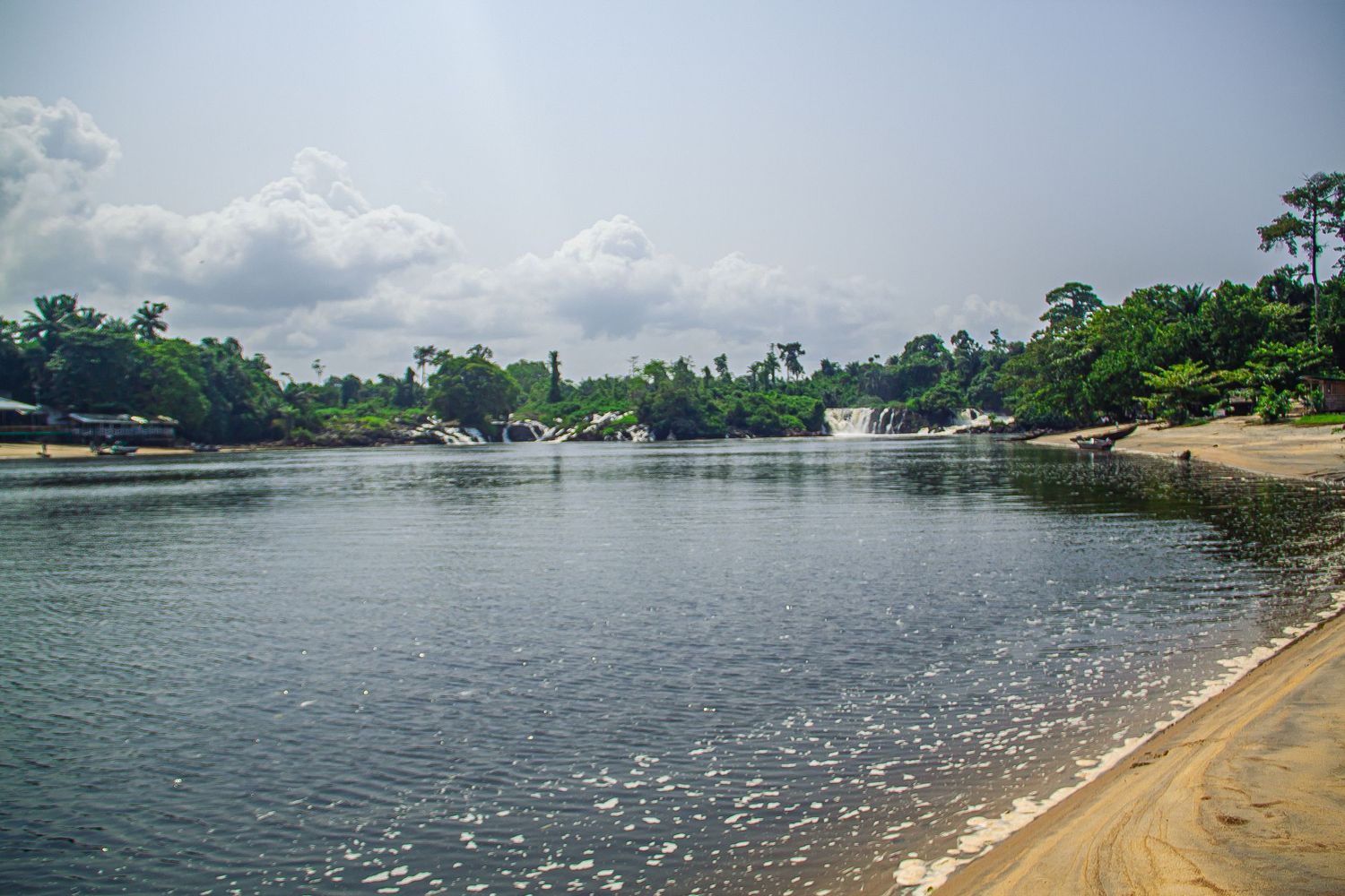 Peaceful sandy beach beside a wide calm river with waterfalls and tropical forest backdrop.