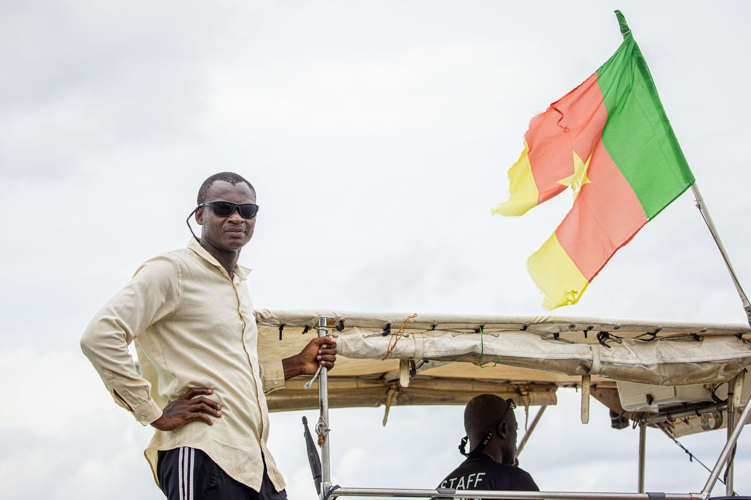 Man in white shirt standing near a Cameroon flag on a boat under a cloudy sky.