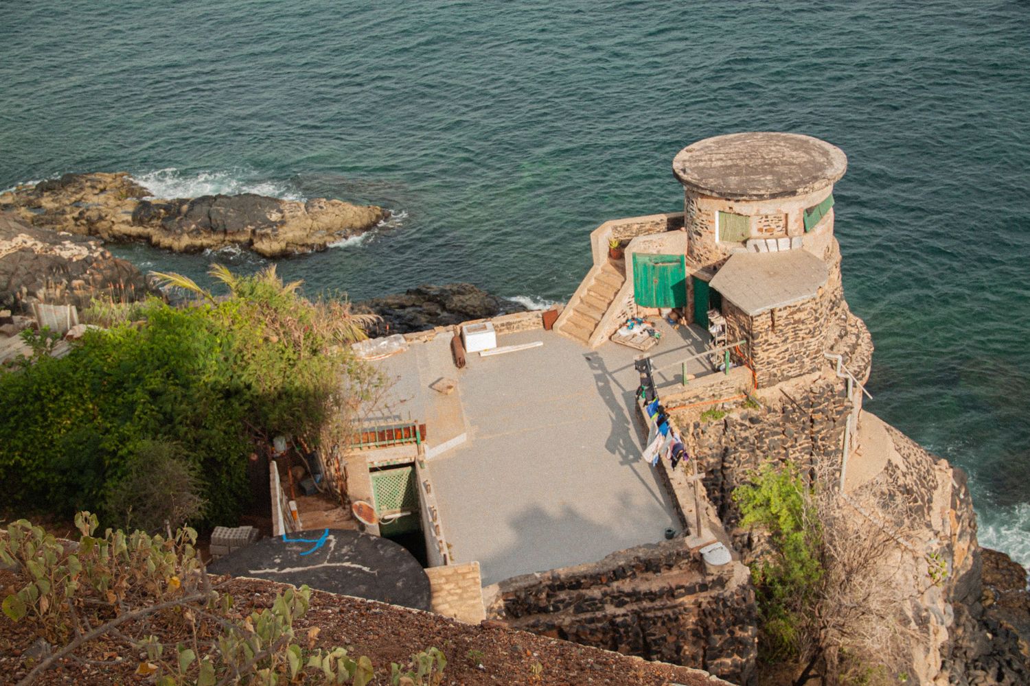 Aerial view of a historic stone fortress with a round tower and green doors perched on rocky cliffs above turquoise ocean waters.