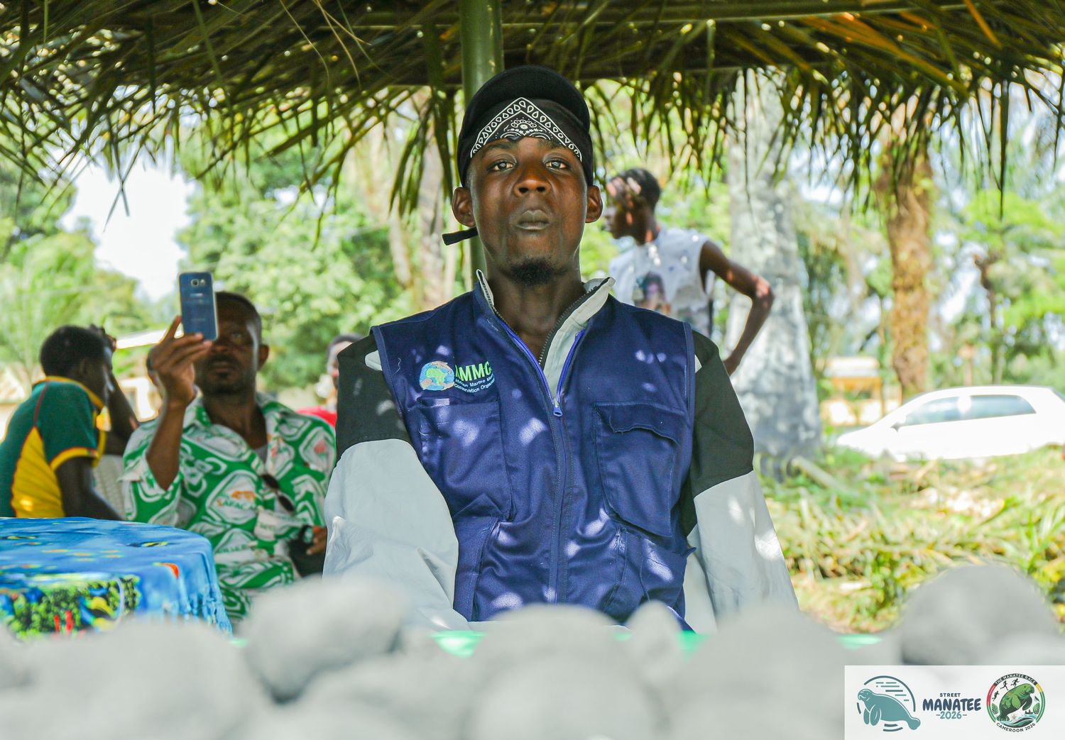 A man wearing a blue vest stands outdoors under a thatched shelter with white objects on a table.