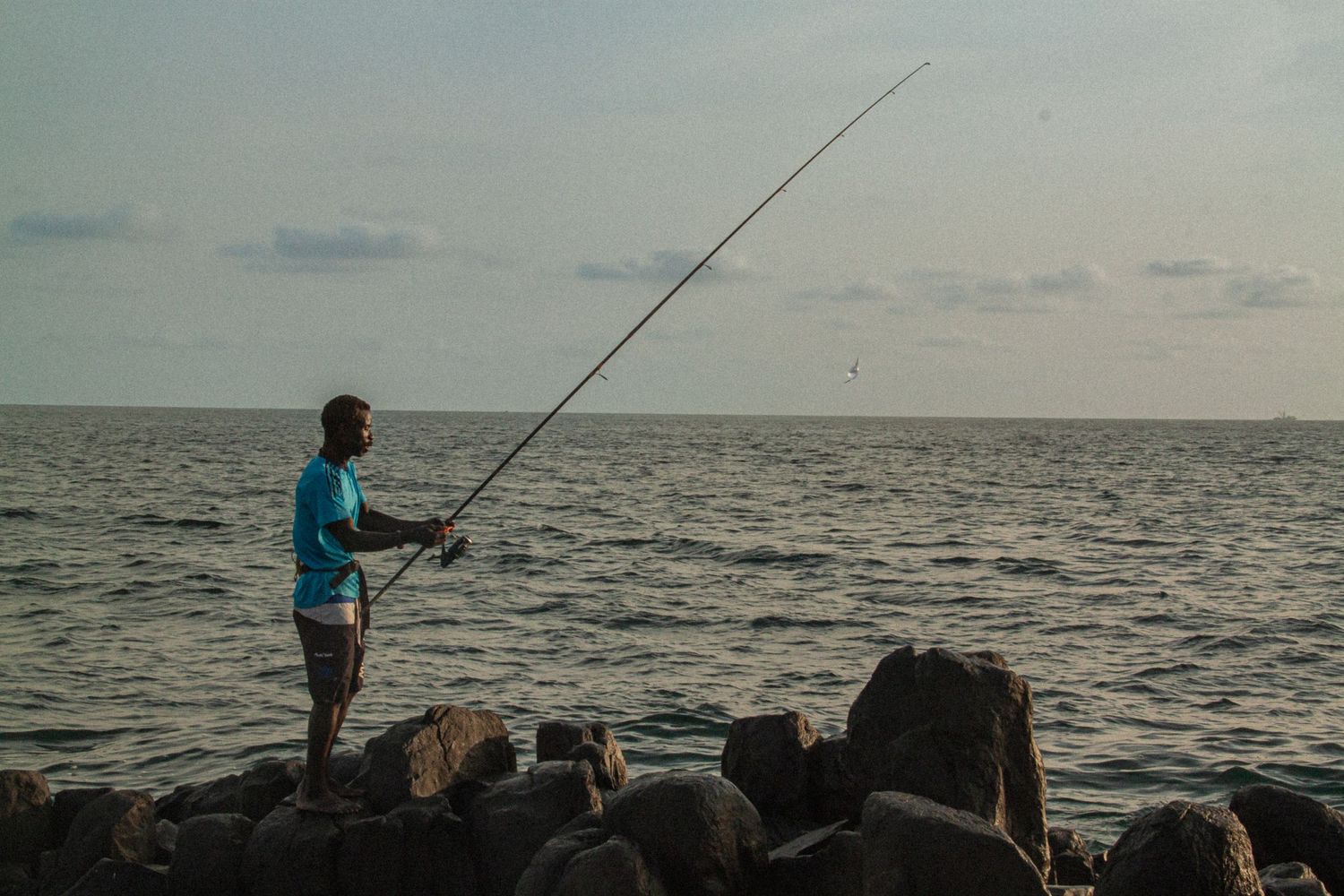 A man fishing with a long rod standing on rocky shoreline with calm ocean waters at sunset.