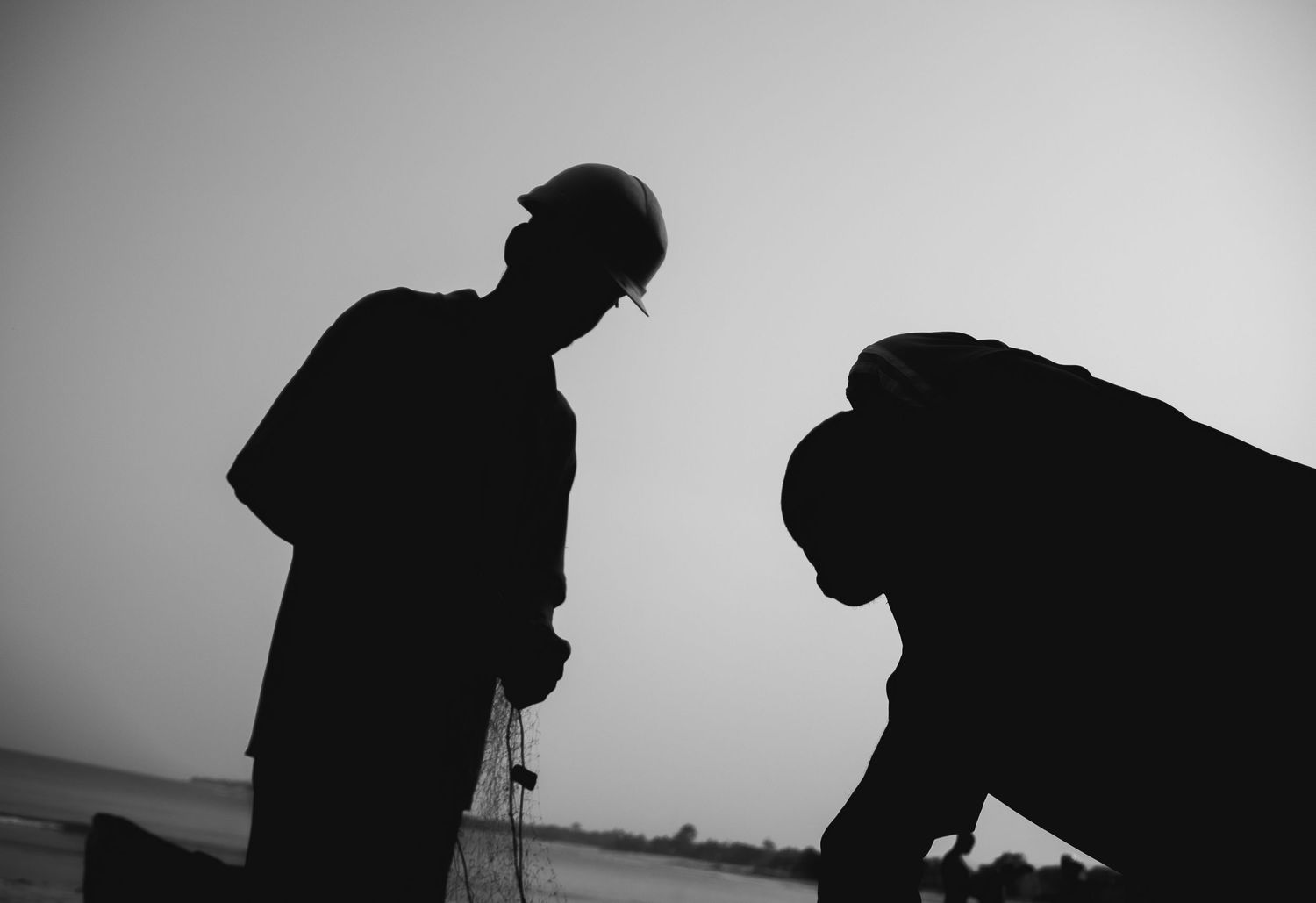 Black and white silhouette of two construction workers wearing hard hats working outdoors.