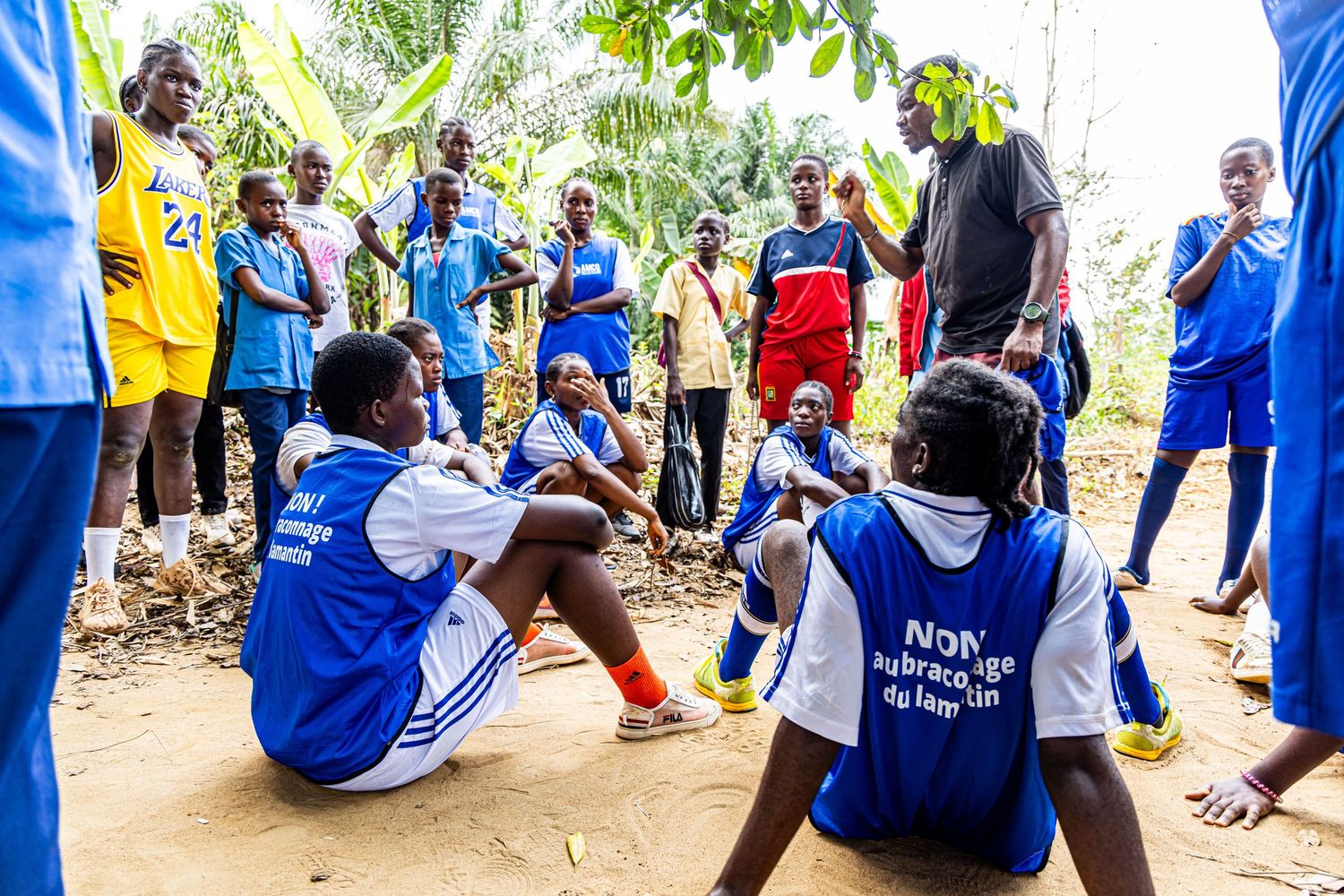 A group of athletes in blue vests sit and stand together on a dirt ground during a team huddle.