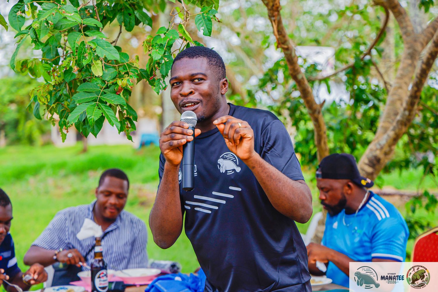 Energetic man speaking into microphone at outdoor community gathering under tree.