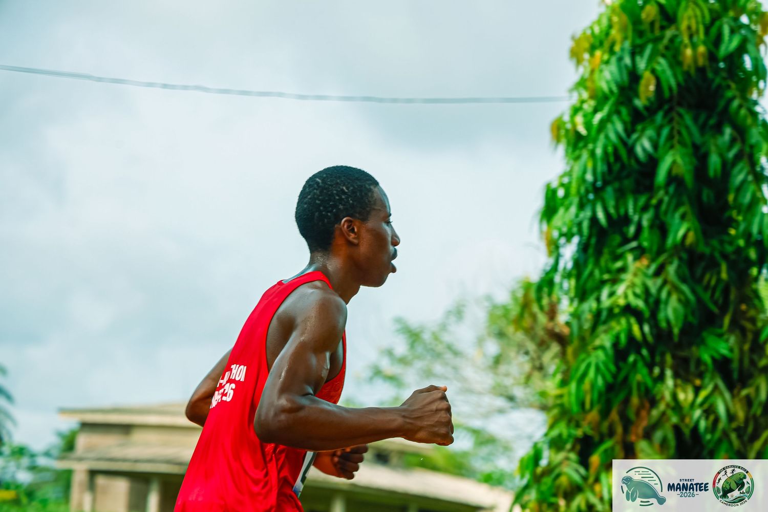 A young male athlete in a red tank top runs outdoors near tropical trees.