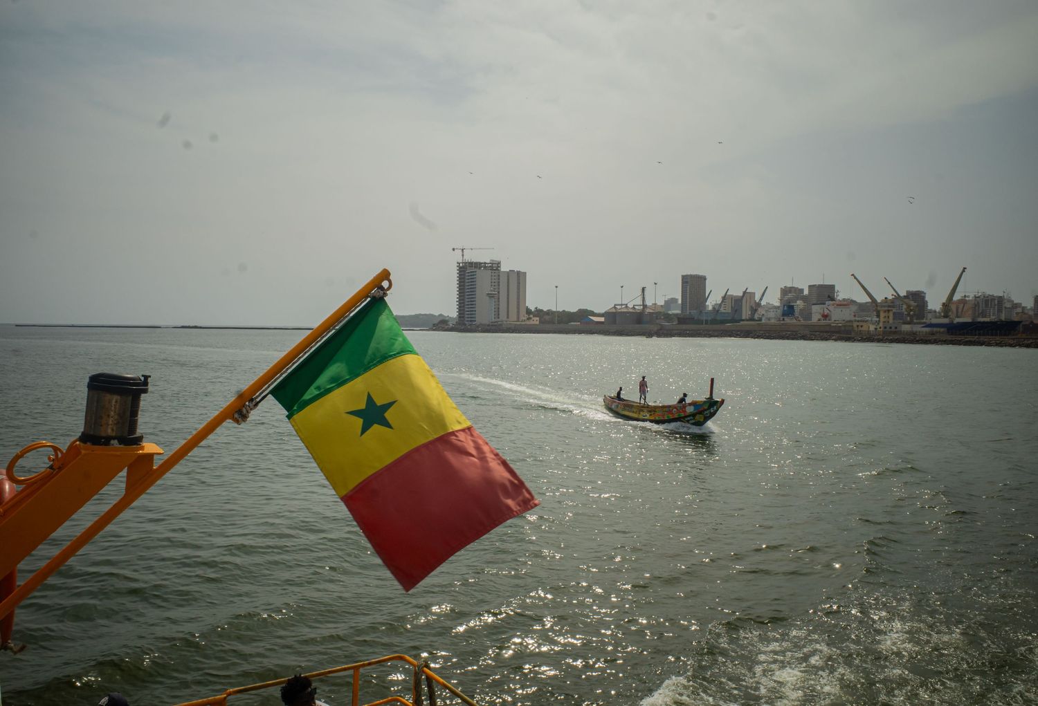 Senegal flag waving on a boat with a wooden fishing vessel and Dakar skyline behind.