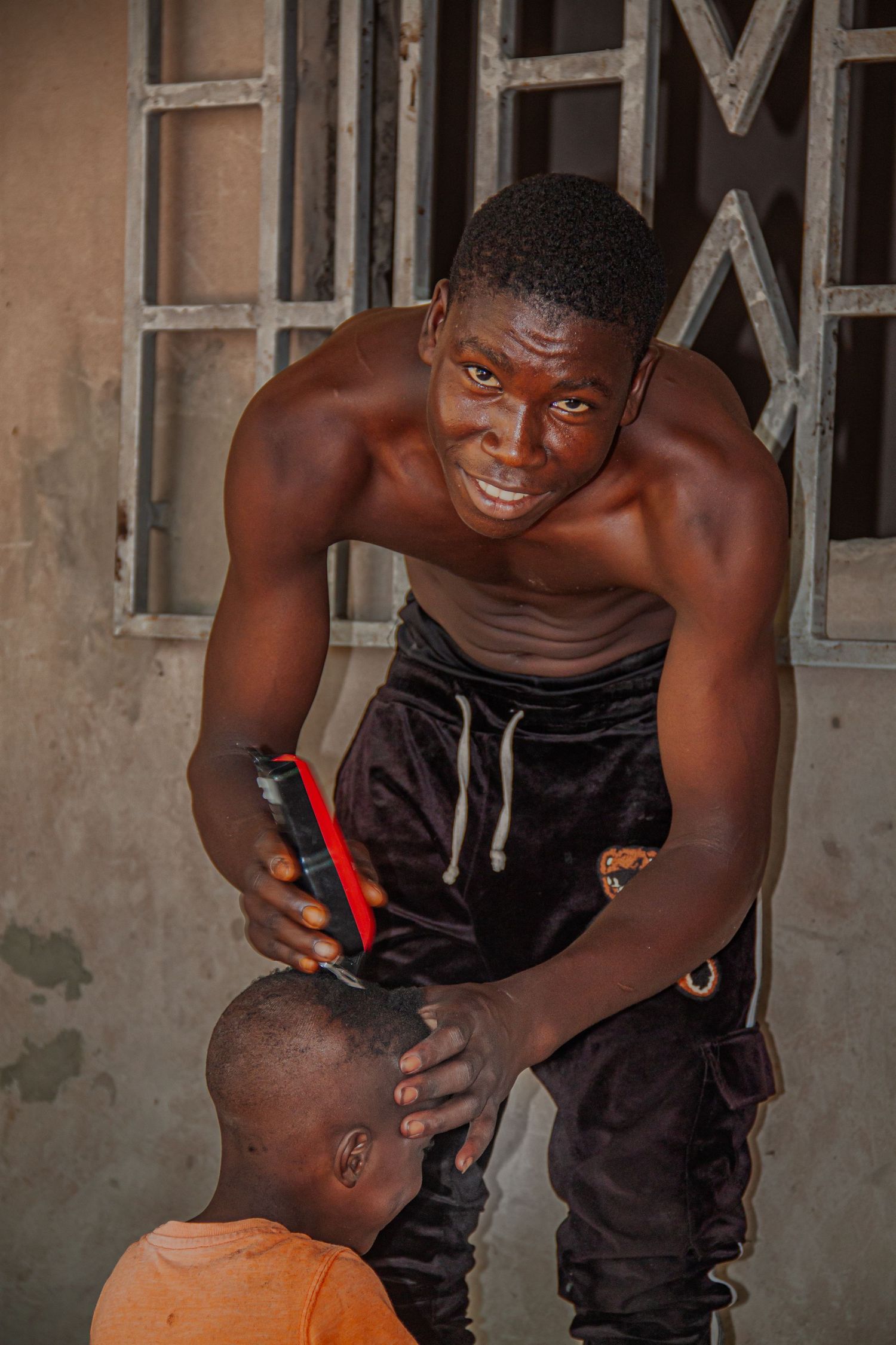 Shirtless young man cutting a childs hair with electric clippers outside a building in Senegal.