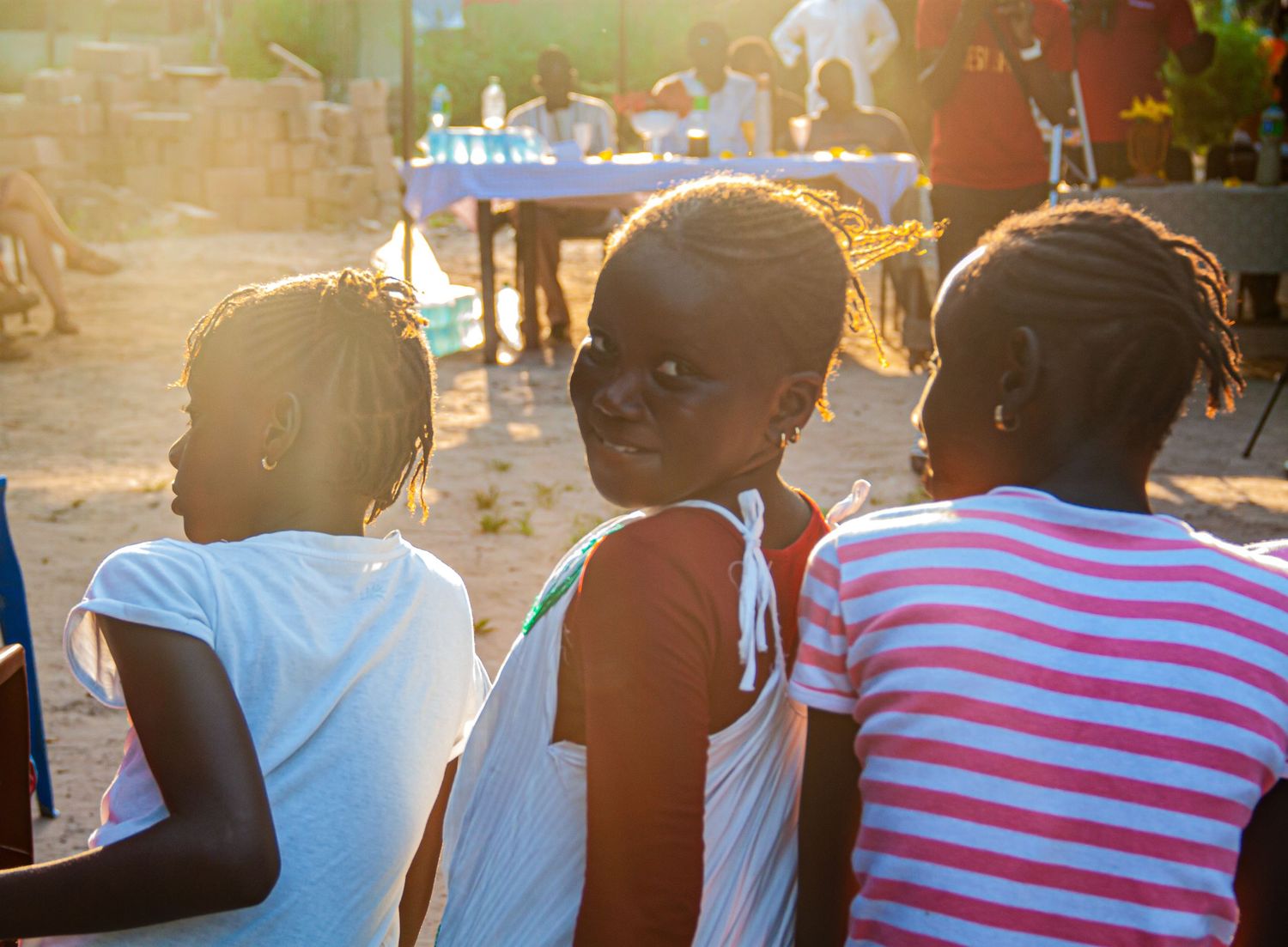 Three young African girls sitting together outdoors at a sunset community gathering event.