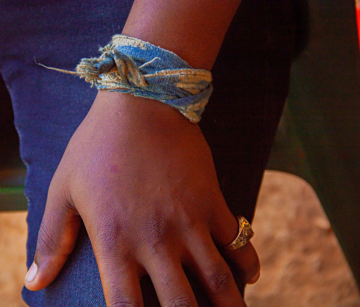 Close-up of African person's hand wearing blue fabric bracelet and gold ring against red dirt background.