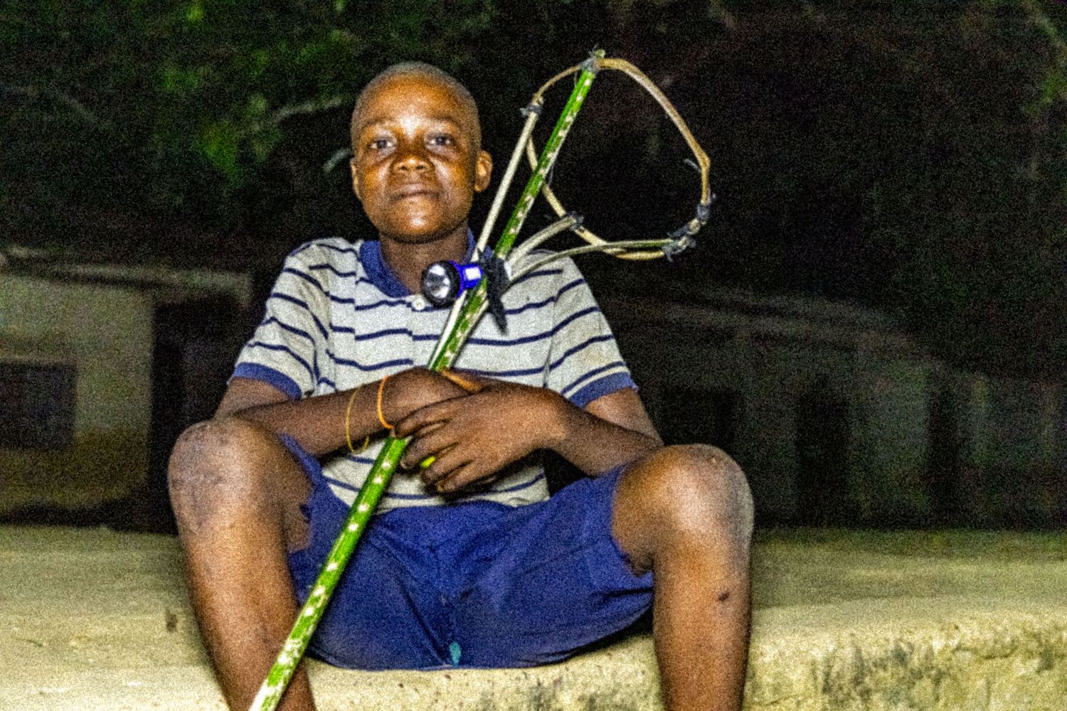 Young African boy sitting outdoors at night holding a handmade bamboo toy racket.