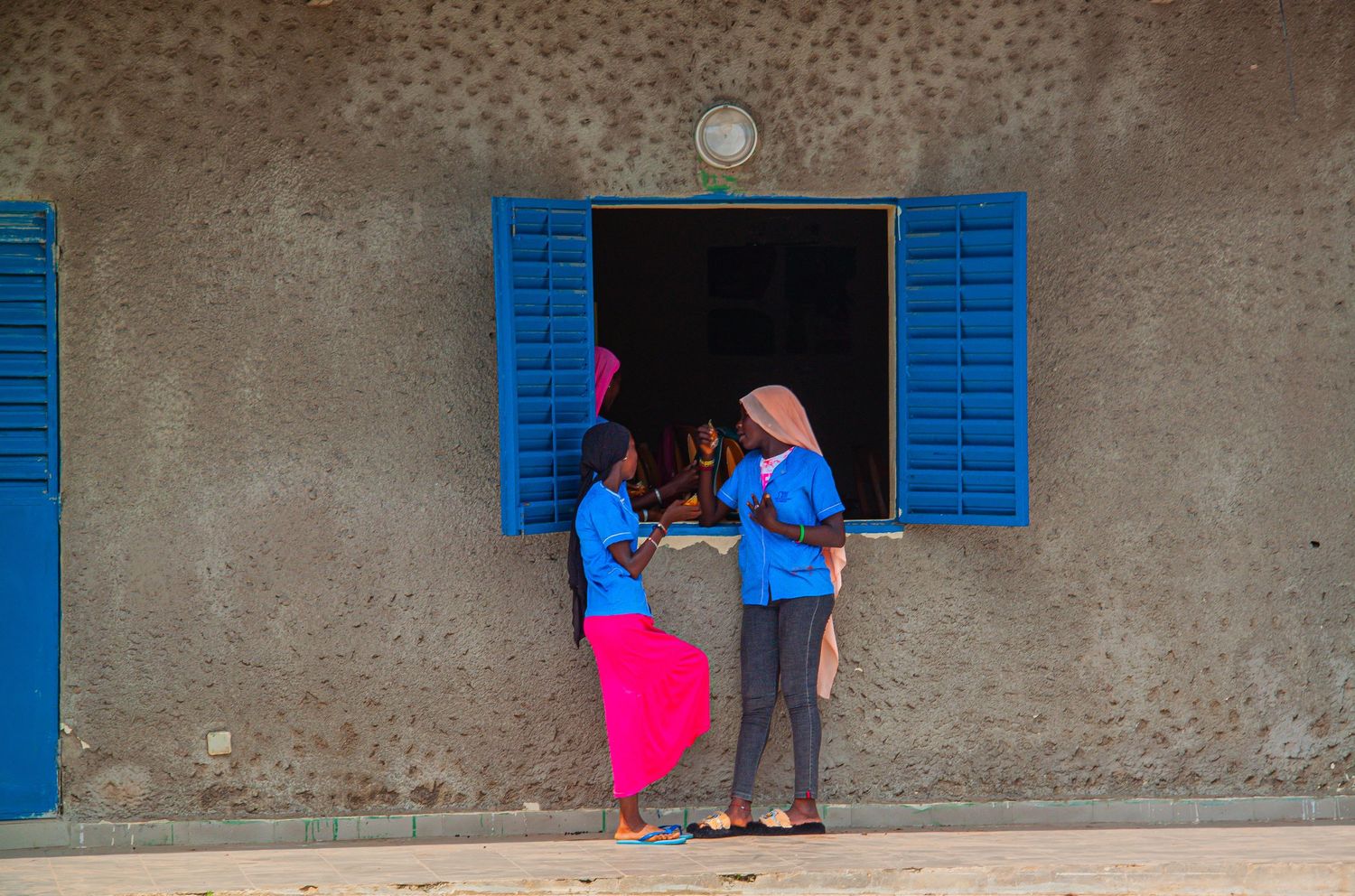 Two African girls in blue uniforms conversing beside a building with blue shuttered window.