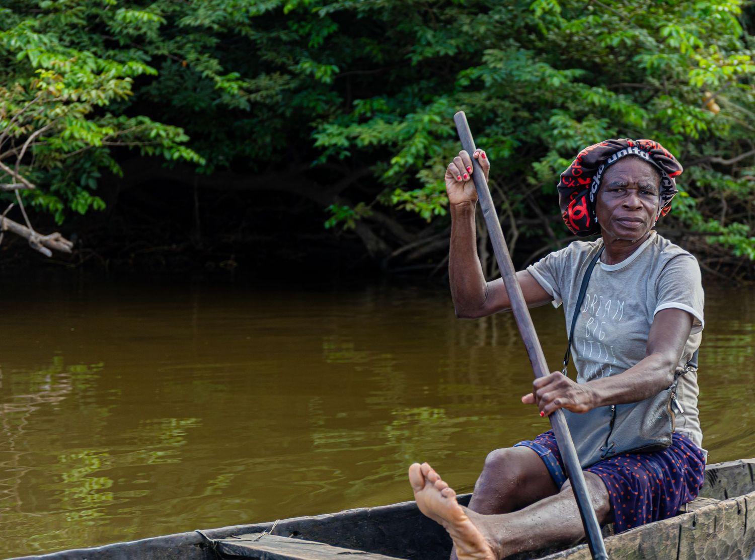 African woman paddling a wooden canoe through a calm brown river surrounded by dense jungle.