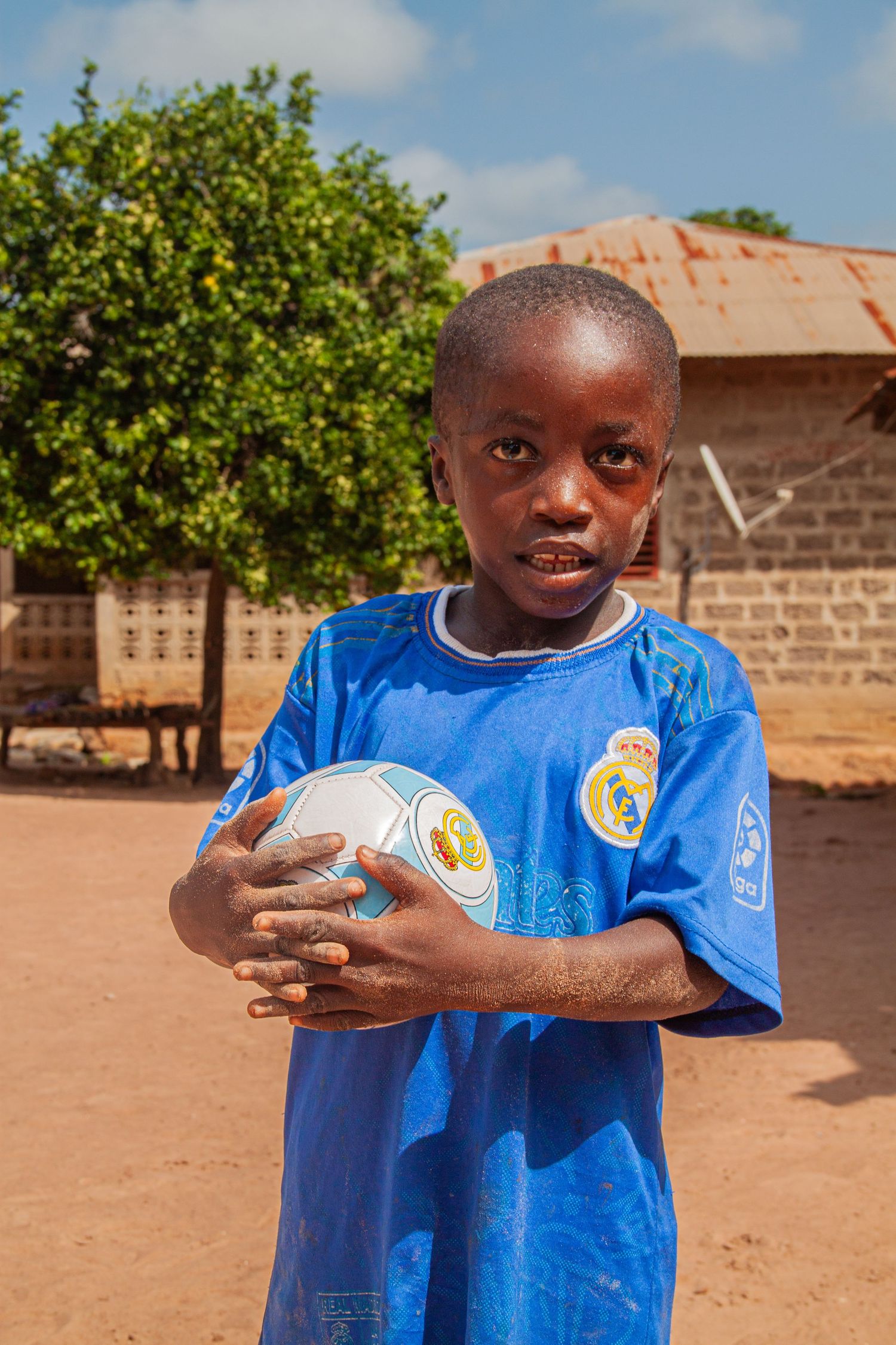 Young African boy in a blue Real Madrid jersey holding a soccer ball outdoors in a rural village setting.